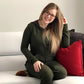 A woman is sitting on a couch wearing a dark green DIBY Club Gabriela Onsie Pajama that features the hood and kangaroo pocket pouch.