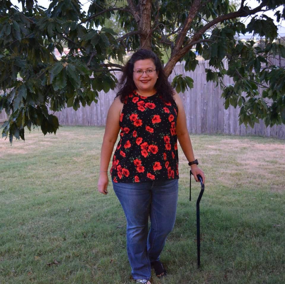 A photo of a woman in a yard wearing the Althea Racerback Tank Top on a black knit with red poppies and white dots. She is standing next to a tree, holding a cane.