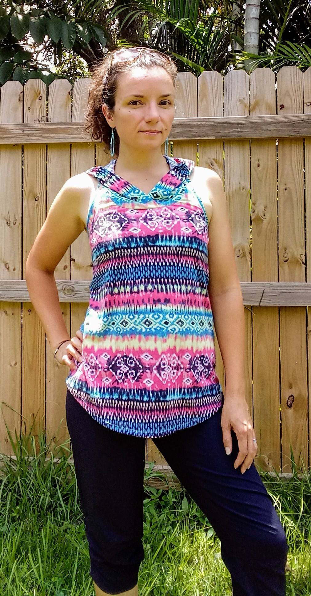 Photo of woman in a backyard wearing an Althea Racerback Tank Top - the tank is made from a knit fabric with a blue, pink, and purple pattern. She is wearing the hooded version with the hood down.