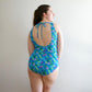 Back view of woman wearing mermaid-scaled blue, green, and purple DIBY Club Amelia one-piece swimsuit with back tie.