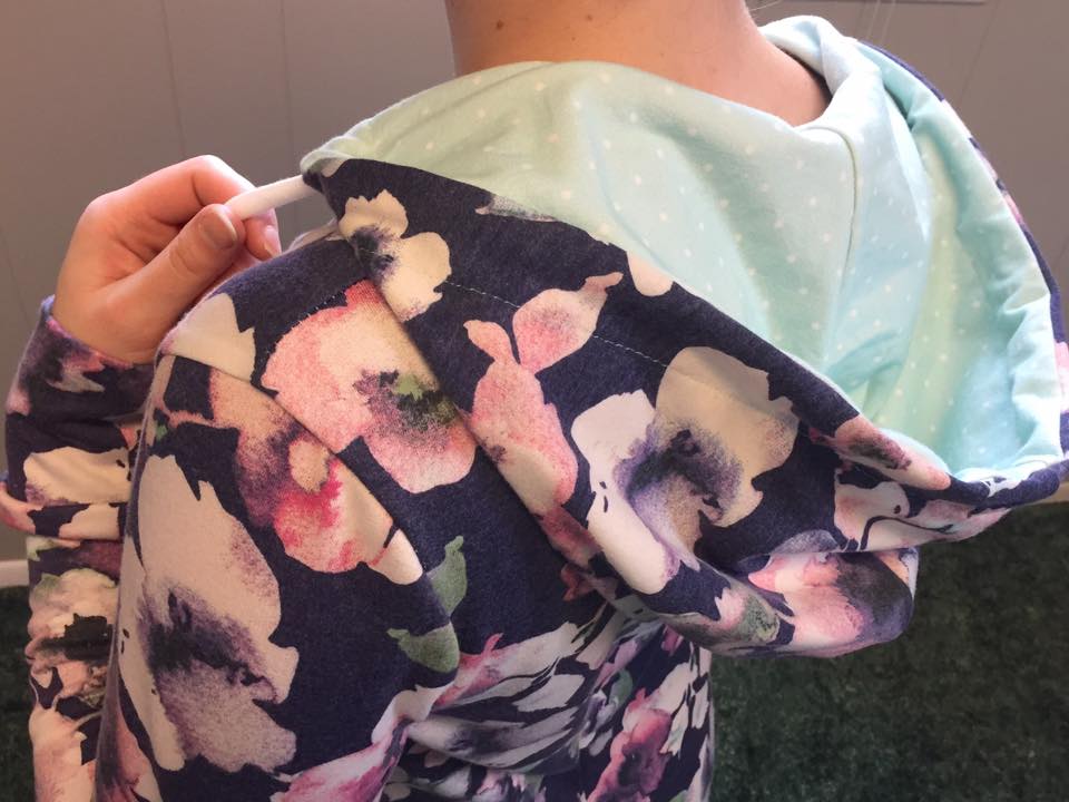 A close up of a person wearing a hooded DIBY Club Gabriela Onsie with a drawstring. The Gabriela is made with large floral knit with a dark blue background. The hood is liked with a light blue knit with white polka dots.