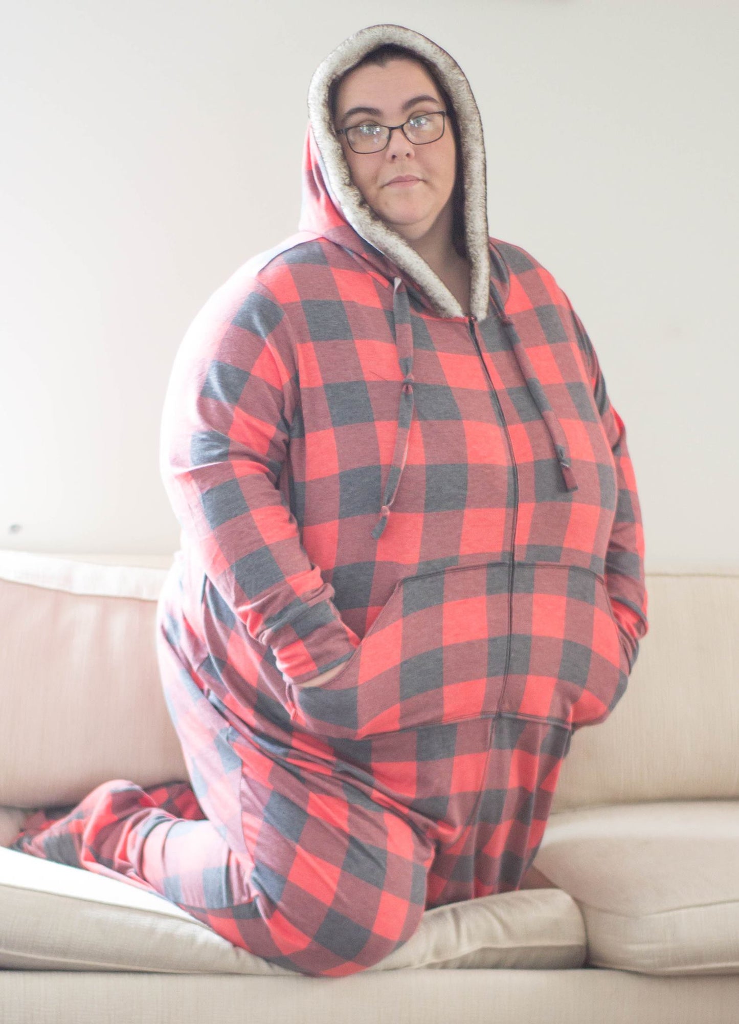 A woman kneeling on a couch wearing a footed, red buffalo plaid DIBY Club Gabriela Onsie. The hoodie is lined with thick "sherpa" style fleece.