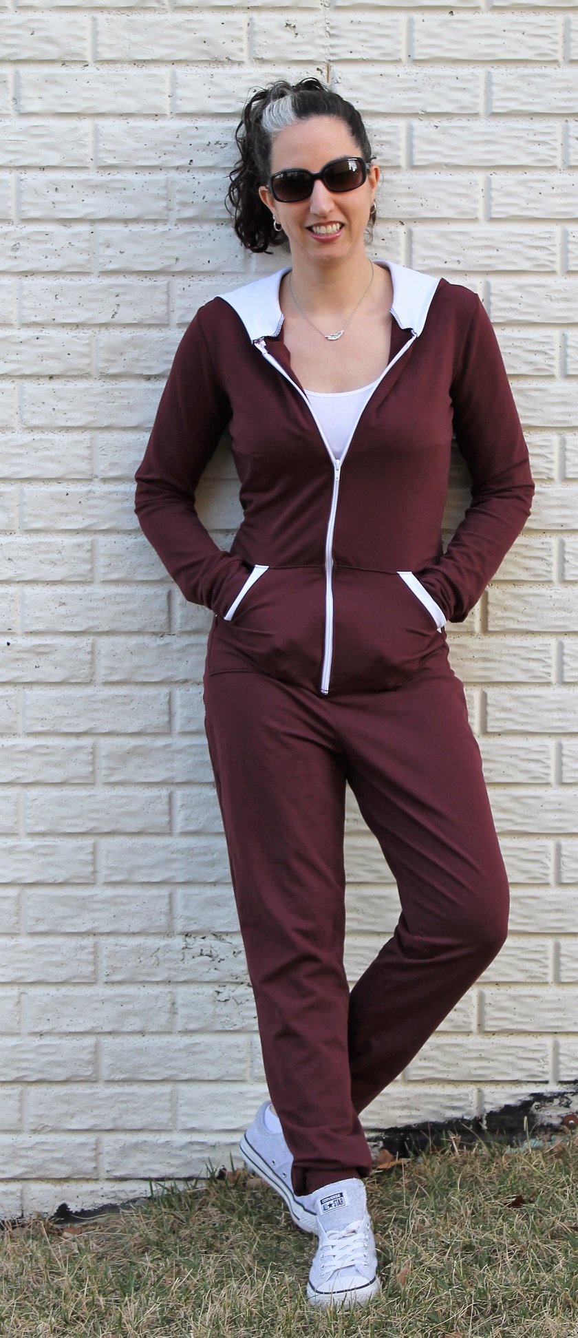 A woman leaning against a wall wearing the DIBY Club Gabriela Onsie. The maroon Gabriela has a white zipper, white pocket trim, and a white lining in the hood. The Gabriela is styled as a sporty jumpsuit.