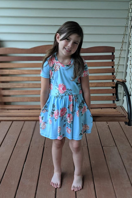 A young girl is wearing the Samantha Gathered Dress pattern with short sleeves and knee length option. The fabric is a pink floral on light blue. 