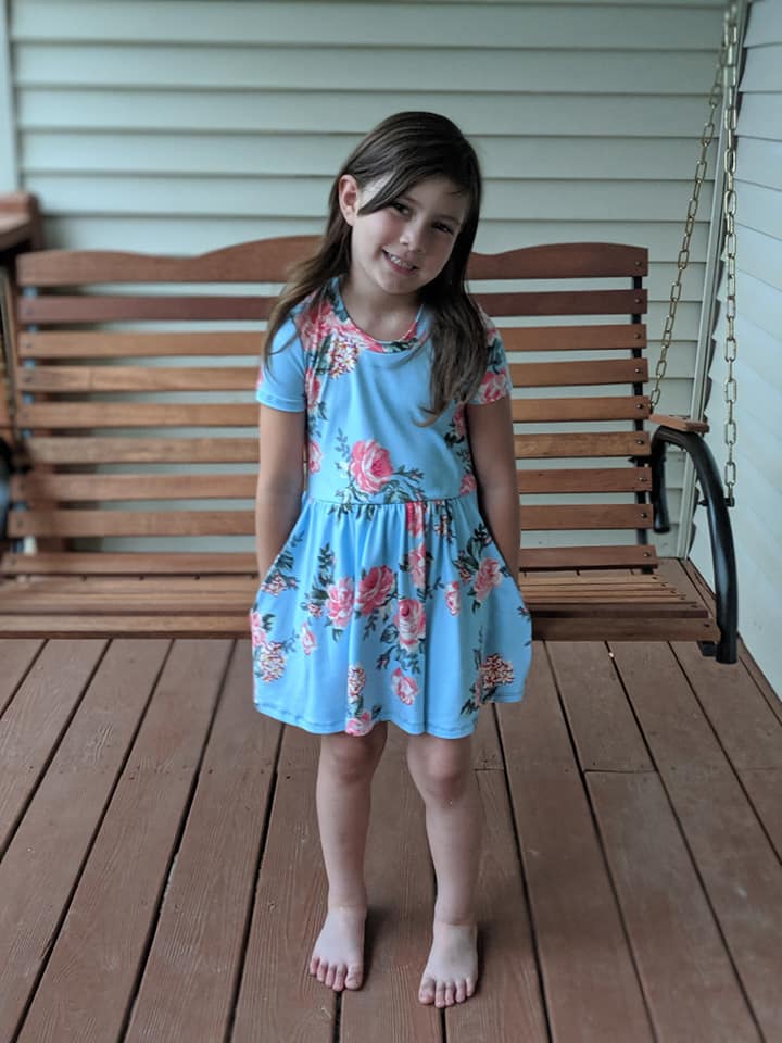 A young girl is wearing the Samantha Gathered Dress pattern with short sleeves and knee length option. The fabric is a pink floral on light blue. 