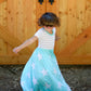 A young girl twirls in her maxi-length, short sleeve version of the Samantha Gathered dress.  The top of the dress is a white and gray striped pattern, and the skirt is light blue with large white stars. 