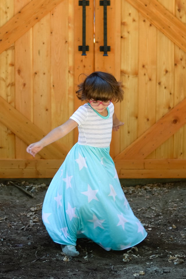 A young girl twirls in her maxi-length, short sleeve version of the Samantha Gathered dress.  The top of the dress is a white and gray striped pattern, and the skirt is light blue with large white stars. 