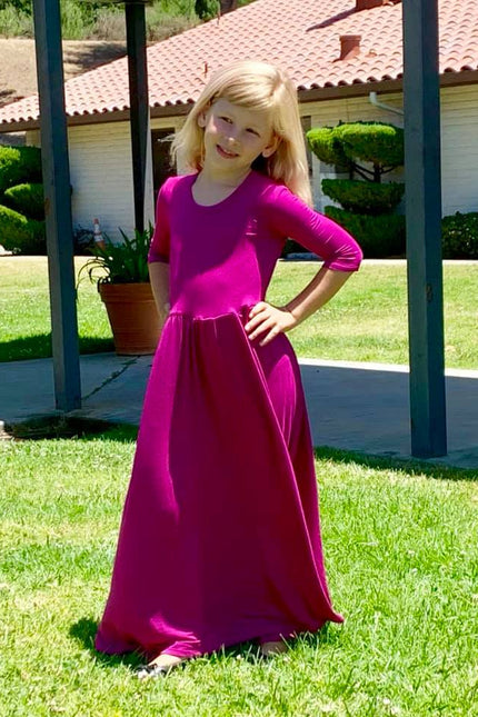 A young girl is wearing a fuchsia, three quarter length sleeve, maxi length Samantha Gathered Dress. 