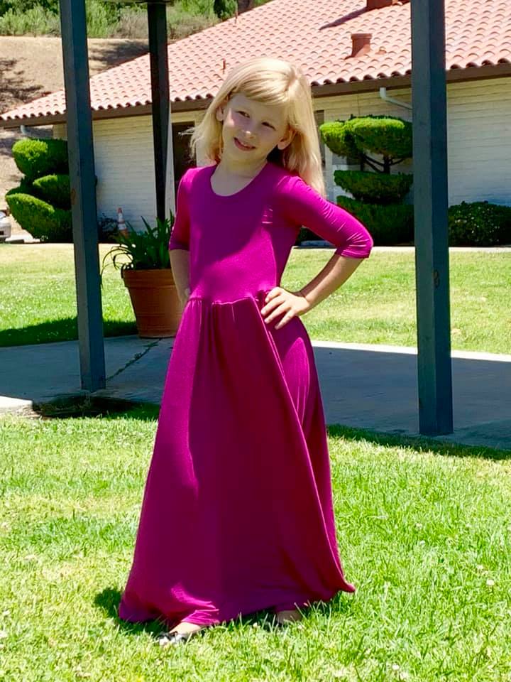 A young girl is wearing a fuchsia, three quarter length sleeve, maxi length Samantha Gathered Dress. 