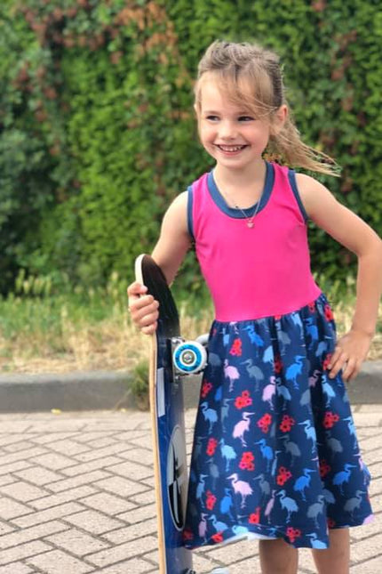A young girl holds her skate board while waring a short sleeve, knee length Samantha Gathered Dress. The top of the dress is pink, trimmed with blue and the skirt is blue with birds. 