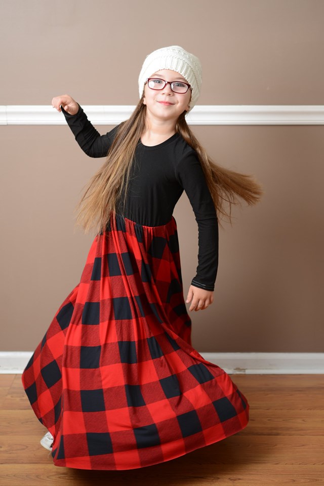 A young girl is wearing the Samantha gathered dress pattern with long sleeves and maxi length. The top is black and the bottom is a red and black gingham pattern. 