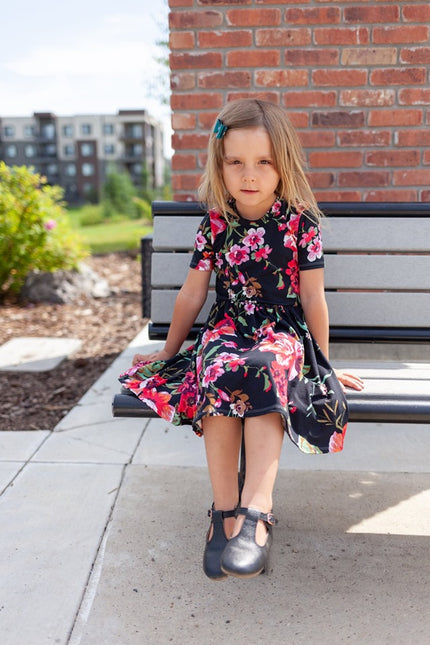 A young girl sits on a bench while wearing a floral themed, short-sleeved, knee length Samantha Gathered Dress. 