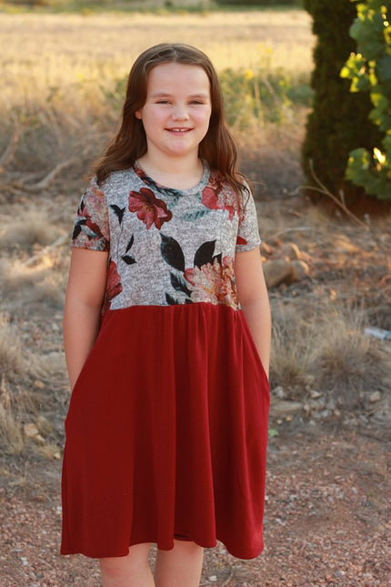 A young girl wears a short sleeve, knee length version of the Samantha dress pattern. The top is a floral design and the skirt is red. 
