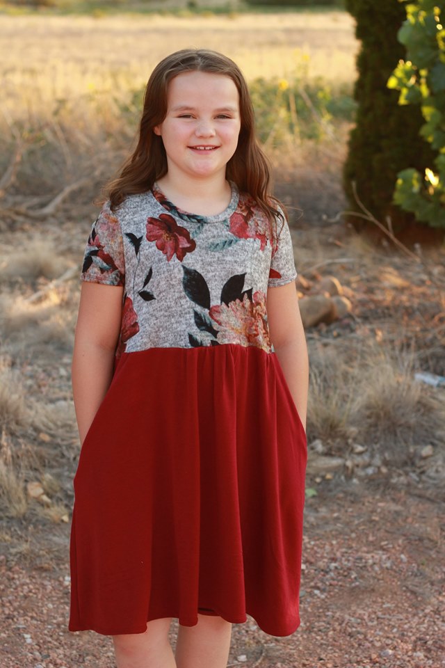 A young girl wears a short sleeve, knee length version of the Samantha dress pattern. The top is a floral design and the skirt is red. 